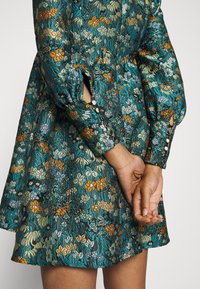 Kurzärmliges Kleid in Petrol mit floralen Mustern in Orange, Gelb und Blau. Verfügt über strukturierten Stoff, Knopfakzente und ausgeschnittene Ärmel.