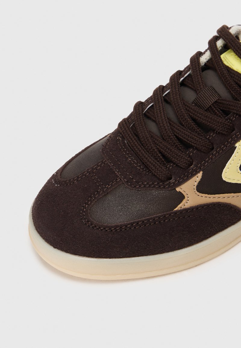 Baskets en suède marron avec un bout en cuir lisse marron et des accents jaunes, dotées de lacets noirs et d'une semelle en caoutchouc neutre.