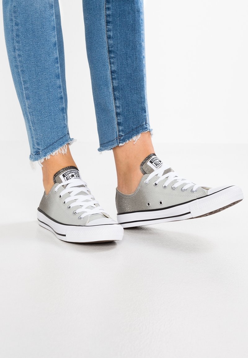 Silver canvas sneakers with white laces, black outsole, and logo accents, paired with frayed-edge blue jeans.