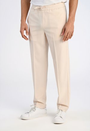 Pantaloni beige con vita elasticizzata e vestibilità morbida, dotati di tasche laterali e dettagli a piega, abbinati a sneakers bianche.