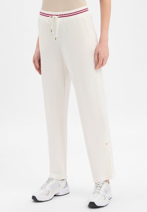 Kobieta ubrana w kremowe spodnie dresowe z czerwoną, pasiastą elastyczną talią i białe trampki, stojąca na tle jednolitego białego tła.