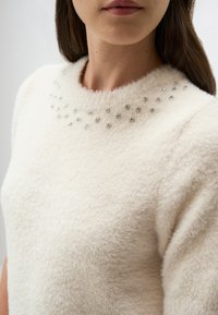 Pull en crème avec une texture douce et duveteuse et un col rond, orné d'accents en strass disposés en motif circulaire.