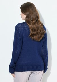 Femme aux longs cheveux bruns ondulés, portant un pull en maille bleu marine et un pantalon clair, tournée légèrement loin de l'appareil.