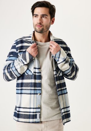Flannel shirt in navy, light blue, and white plaid. Features buttons, a collar, and long sleeves. Worn over a light gray t-shirt.
