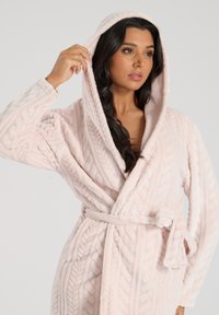 Accappatoio rosa chiaro con cappuccio realizzato in tessuto morbido e testurizzato con un motivo a trecce. Presenta una cintura in vita e tasche visibili.