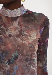 Haut transparent foncé avec des motifs abstraits tourbillonnants multicolores, comprenant des touches de rouge, bleu, violet et vert, porté par une personne avec des boucles d'oreilles anneaux en or.