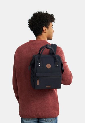 Navy backpack with a textured finish, top handles, padded shoulder strap, front zip pocket, and a circular logo accent.