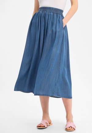 Vrouw draagt een blauwe denim midi-rok met een elastische taille en zijzakken, gecombineerd met roze open-toe platte sandalen.