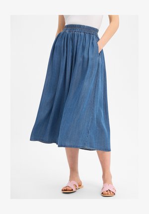 Vrouw draagt een blauwe denim midi-rok met een elastische taille en zijzakken, gecombineerd met roze open-toe platte sandalen.