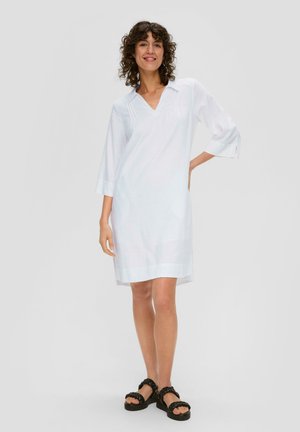 Weißes Leinenkleid mit V-Ausschnitt, dreiviertelärmel, seitlichen Schlitzern und lockerer Passform, kombiniert mit schwarzen geflochtenen Sandalen.