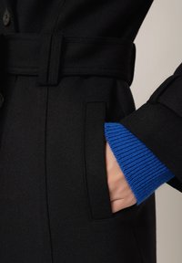 Czarny płaszcz z paskiem w talii i bocznymi kieszeniami, z niebieskim prążkowanym mankietem widocznym od wewnętrznej strony rękawa. Gładka tekstura.