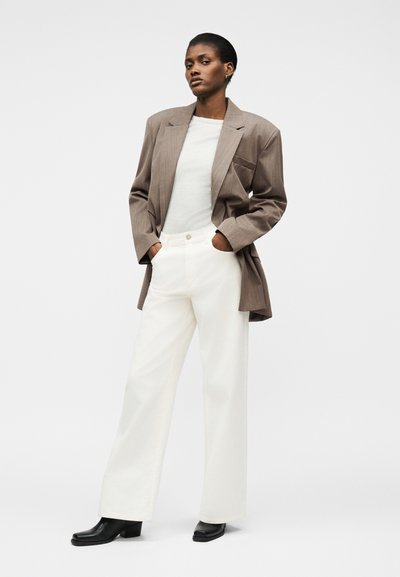 Γυναίκα που στέκεται με τα χέρια στις τσέπες, φορώντας καφέ oversized σακάκι, λευκό τοπ, λευκά παντελόνια με φαρδιά μπατζάκια και μαύρα μποτάκια μέχρι τον αστράγαλο, πάνω σε απλό φόντο.