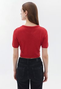 Maglione rosso corto con maniche corte, tessuto testurizzato e scollatura ampia, abbinato a jeans neri a vita alta con tasche posteriori.
