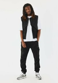 Sort ærmeløs vest over en hvid t-shirt, kombineret med sorte cargo-bukser med lommer og hvid-sorte sneakers.