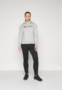 Pilkas hoodie su tamsiai mėlynu logotipu, raišteliais ir ribotais rankogaliais, derinamas su juodomis lieknomis kelnėmis ir juodais sportbačiais neutraliame fone.