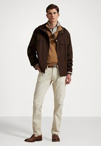 Καφέ ελαφρύ μπουφάν πάνω από ένα μπεζ πουλόβερ, σε συνδυασμό με ανοιχτόχρωμο τζιν και καφέ δερμάτινα παπούτσια. Περιλαμβάνει πουκάμισο με καρό σχέδιο από κάτω.