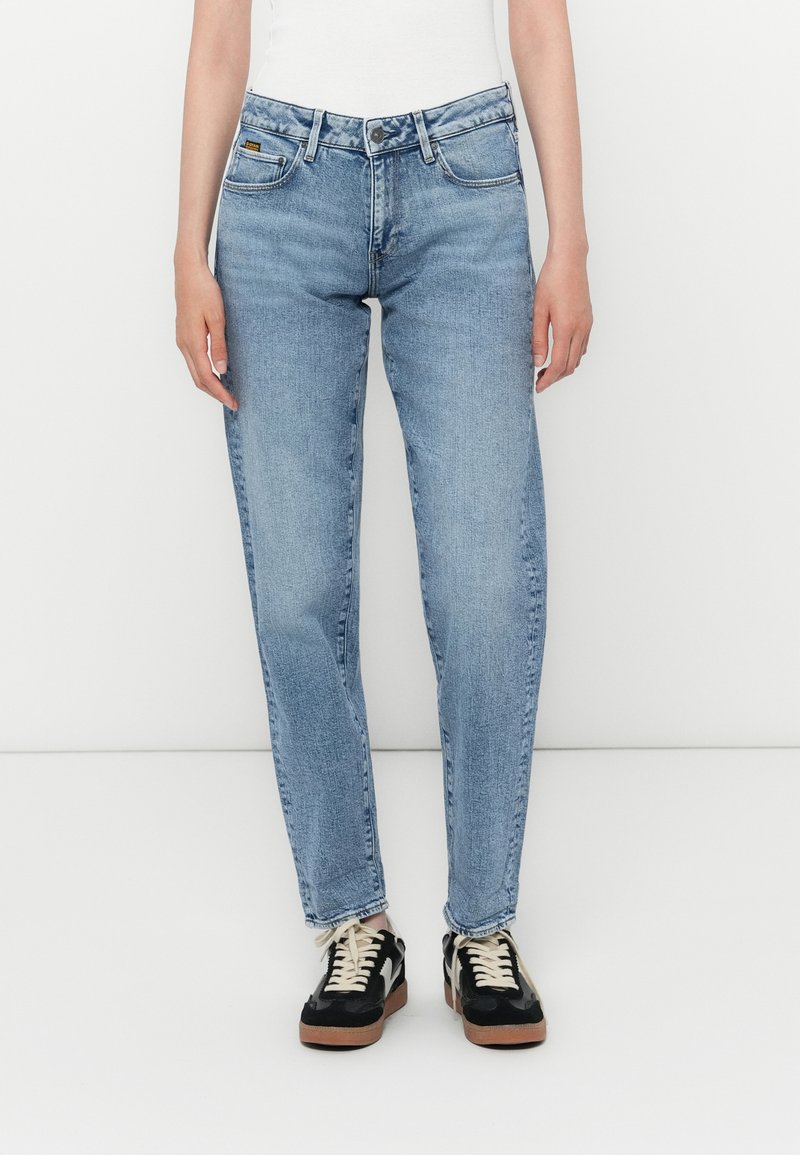 Ανοιχτό μπλε τζιν παντελόνι με χαλαρή εφαρμογή, πέντε τσέπες και διακριτική φθορά. Συνδυασμένο με μαύρα πάνινα παπούτσια με λευκές λεπτομέρειες.