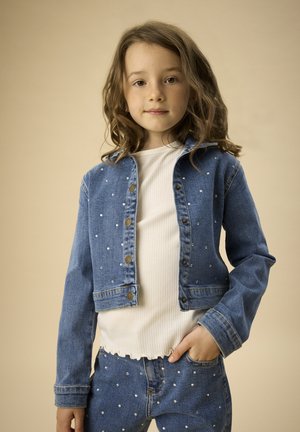 Veste en jean avec détails en perles, fermeture par boutons et manches longues. Portée avec un haut blanc côtelé et un jean, affichant un look décontracté.