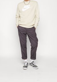 Person som bär en beige Nike-tröja, mörkgrå upprullade byxor, vita strumpor, svartvita sneakers och ett silverfärgat kedjehalsband.