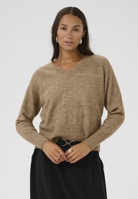 Pull en V à texture marron avec de longues manches et un ourlet côtelé, porté sur un pantalon noir taille haute, doté d'une ceinture avec une quincaillerie circulaire.