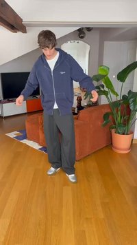 Navy zip-up hoodie with white logo, paired with gray wide-leg pants and silver sneakers; wooden floor and indoor plants in background.