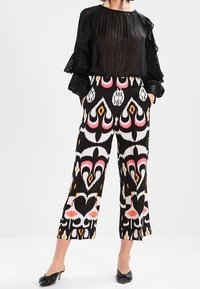 Femme portant un chemisier noir transparent à manches longues et un pantalon taille haute à jambes larges avec des motifs géométriques audacieux en noir, blanc, rose et orange.