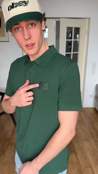 Νεαρός άνδρας που φοράει πράσινο πόλο πουκάμισο και λευκό-πράσινο καπέλο δείχνει προς ένα λογότυπο στο στήθος του πουκάμισου σε δωμάτιο με ξύλινο πάτωμα.