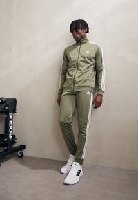 Olivovo zelený teplákový komplet s bielymi bočnými prúžkami, ktorý obsahuje bundu so zipsom a zúžené nohavice. Komplet doplnený o biele tenisky. Jednoduchá textúra látky.