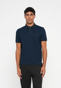 A camada de polo azul-marinho possui um tecido texturizado, mangas curtas, um colarinho riscado e um pequeno logótipo no peito. Usado com calças pretas.