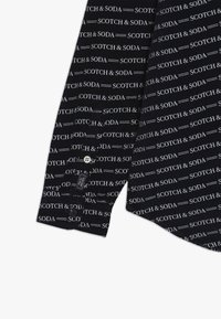 Chemise noire avec un motif répété de texte blanc "Scotch & Soda". Le tissu a une texture lisse, avec une fermeture à bouton au niveau des poignets.