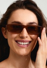 Gafas de sol rectangulares marrones con montura transparente, acabado brillante, usadas por una mujer que toca la montura, con cabello largo y ondulado.