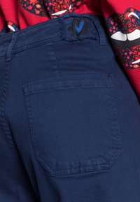 Calças de ganga azul-marinho com um bolso traseiro, apresentando bordas desfiadas e um pequeno desenho de coração bordado em azul na cintura.