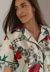 Cămașă cu mânecă scurtă și imprimeu floral în nuanțe de crem, având detalii negre și flori colorate în roșu, albastru și portocaliu, cu accente geometrice.