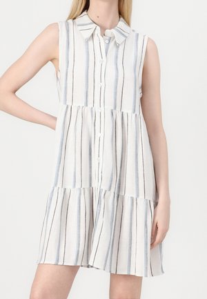 Robe blanche sans manches avec des rayures verticales bleues et noires, boutonnée sur le devant, et une jupe à volants portée par une personne debout.