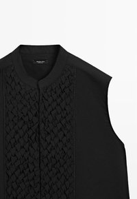 Černý top bez rukávů s kulatým límcem a texturovaným tkaným vzorem táhnoucím se vertikálně středem přední části.