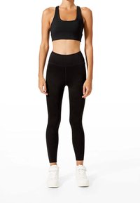 Czarny sportowy biustonosz i legginsy o wysokim stanie wykonane z elastycznego materiału; cechują się bezszwowym designem i gładką, matową teksturą. Białe sneakersy.