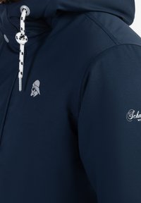Veste bleu marine avec capuche à cordon blanc, logo d'un guerrier casqué sur la poitrine, et broderie du texte « Schm » sur la manche.