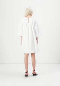 Hvit, kortermet kjole med rund hals og nøkkelhulldetalj på baksiden; brukt sammen med svarte sandaler med stropper. Jevn stofftekstur.