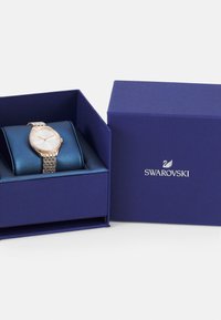 Zegarek Swarovski w granatowym pudełku, z różowo-złotą metalową bransoletą ozdobioną kryształami oraz okrągłą białą tarczą ze złotymi wskazówkami.