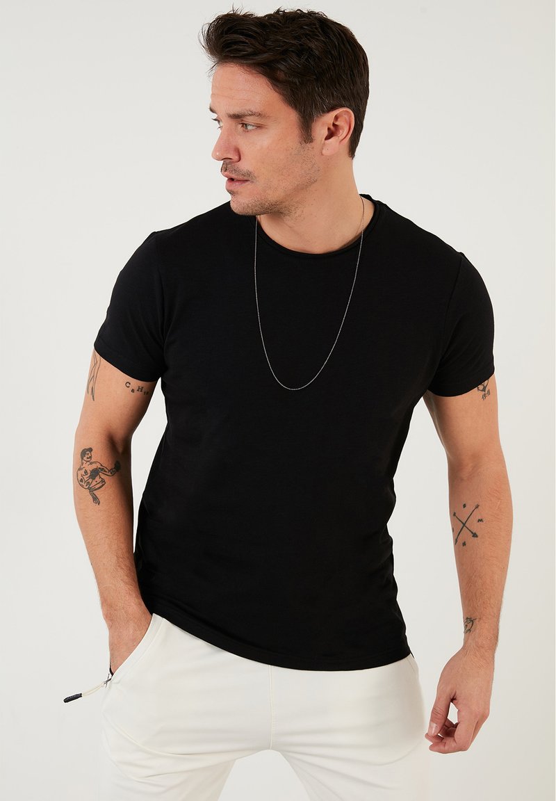 T-shirt nero a maniche corte con scollatura rotonda, realizzato in tessuto leggero. Tatuaggi notevoli sulle braccia, indossato con pantaloni bianchi.