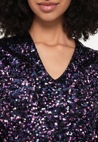 Μαύρο τοπ με ντεκολτέ σε σχήμα V, καλυμμένο με πολύχρωμες παγιέτες (μοβ, ροζ και ασημί) με λαμπερή υφή, που αναδεικνύει ζωντανές χρωματικές διαφοροποιήσεις.