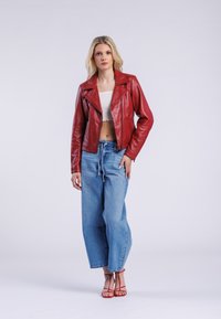 Červená kožená motorkářská bunda s klopovým límcem, v kombinaci s volnými světle modrými džíny. Hladký materiál, přiléhavý střih a zapínání na zip.