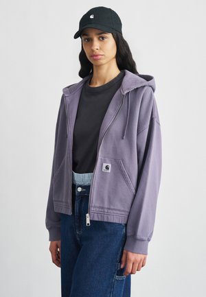 Purple zip-up hoodie with a front pocket and logo patch, worn over a black t-shirt. Paired with high-waisted blue jeans and a black cap.