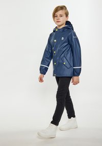 Jūras spēku ūdensnecaurlaidīgs lietusjakas ar kapuci, balti akcenti, dzelteni pogas un sānu kabatas, uzvilkts virs melnām biksēm un baltām sporta apaviem.