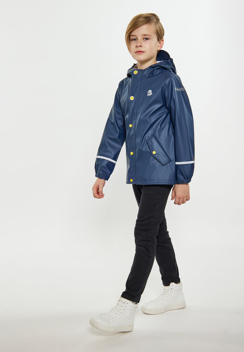 Jūras spēku ūdensnecaurlaidīgs lietusjakas ar kapuci, balti akcenti, dzelteni pogas un sānu kabatas, uzvilkts virs melnām biksēm un baltām sporta apaviem.