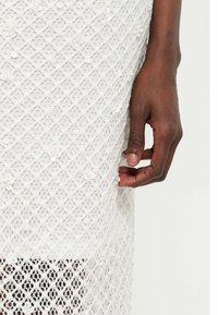 Σκοτεινόχρωμο χέρι με φυσικά νύχια που ακουμπά δίπλα σε λευκό φόρεμα από δαντέλα διακοσμημένο με μικρές λευκές παγιέτες και μοτίβα σε σχήμα ρόμβου.