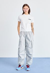 Balti prigludusi marškiniai su "DICKIES" logotipu, šviesiai pilkos kargo kelnės su kišenėmis ir mėlyni sportbačiai su raudonais akcentais. Aiškūs saulėgrąžų akiniai užbaigia vaizdą.