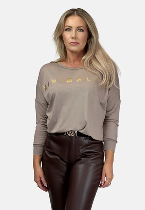 Frau mit langen blonden Haaren, die ein beiges Langarmshirt mit "MINIMALIS" in Gold und dunkellobraunen Lederhosen mit einem schwarzen Gürtel trägt.