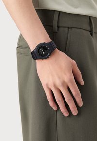 Ceas din rășină neagră cu cadran rotund, bezel texturat, ace în contrast și o curea flexibilă, purtat pe o cămașă gri deschis și pantaloni verzi.