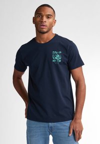 T-shirt en coton bleu marine avec col rond, featuring un design graphique turquoise sur la poche et des manches courtes. Coupe classique.